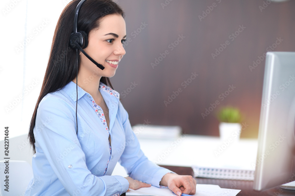Young brunette business woman looks like a student girl working in office. Hispanic or latin american girl talking by headphones at pc Young brunette business woman looks like a student girl working in office. Hispanic or latin american girl talking by headphones at pc