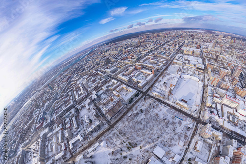 Round panorama aerial view on a winter city under a blue sky with white clouds  rows of streets with high-rise buildings and roofs under snow  a park  trees and a stadium with a football field.