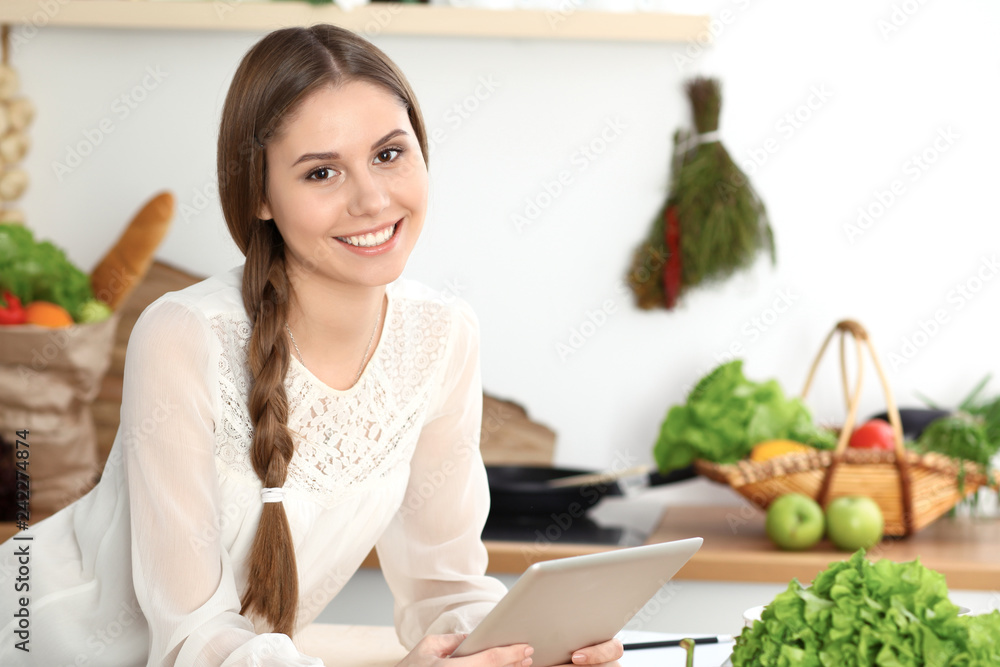 Young woman using tablet while cooking or making online shopping in kitchen. Girl looking at the camera. Healthy lifestyle and vegan meal concepts Young woman using tablet while cooking or making online shopping in kitchen. Girl looking at the camera. Healthy lifestyle and vegan meal concepts