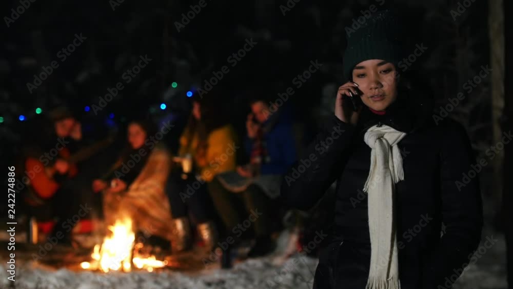 Young woman in black down jacket standing in winter forest and talking on her phone. Group of friends sitting by the fire on the background