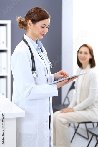 Happy doctor woman at work. Portrait of female physician using tablet computer while standing near reception desk at clinic or emergency hospital. Patient woman sitting at the background. Medicine