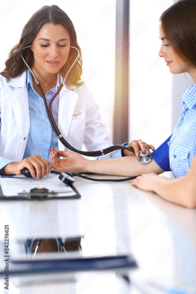 Doctor checking blood pressure of female patient while sitting at the desk in hospital office. Cardiology in medicine and health care concept Doctor checking blood pressure of female patient while sitting at the desk in hospital office. Cardiology in medicine and health care concept