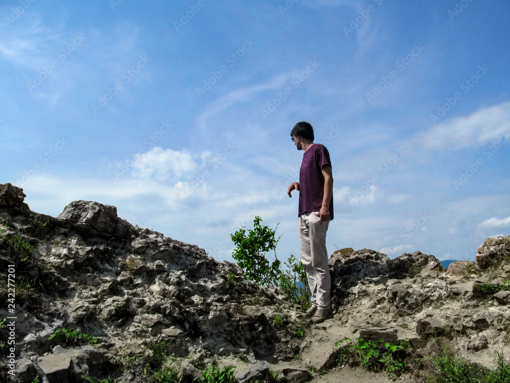 A tall dark-haired young man stands on a rock and looks down - view from the back. Clear sunny day, stone boulders and one adult guy against the sky