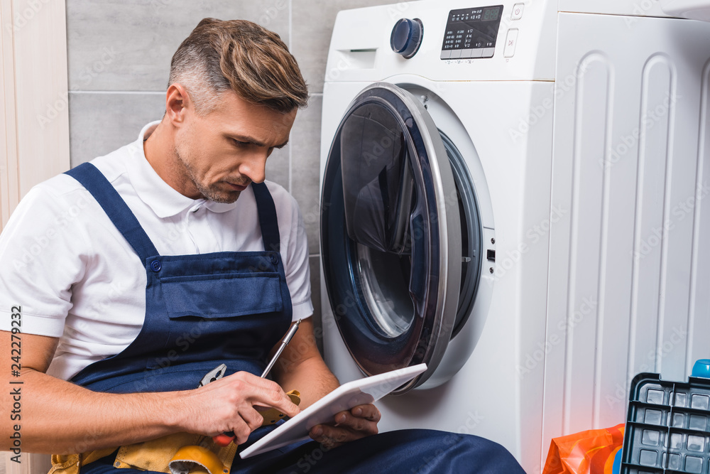 adult repairman holding screwdriver and using digital tablet while repairing washing machine in bathroom adult repairman holding screwdriver and using digital tablet while repairing washing machine in bathroom
