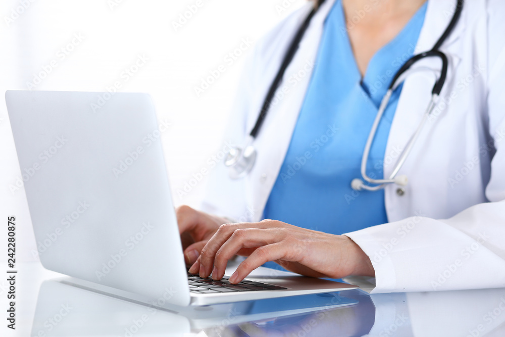 Doctor typing on laptop computer while sitting at the glass desk in hospital office. Physician at work. Medicine and healthcare concept Doctor typing on laptop computer while sitting at the glass desk in hospital office. Physician at work. Medicine and healthcare concept