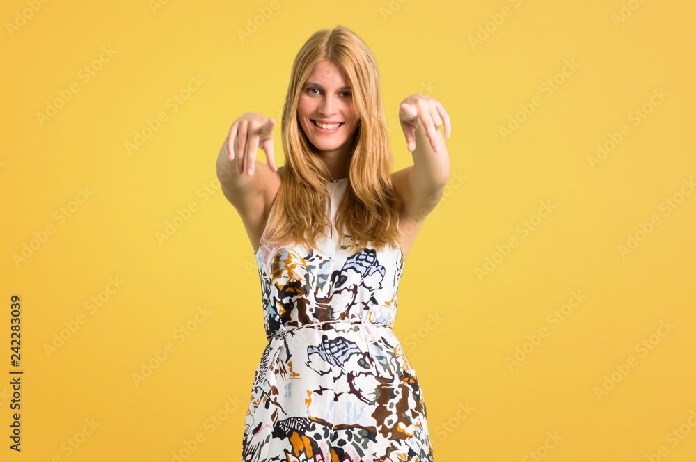 Blonde young girl in a summer dress pointing with finger at someone and laughing a lot on yellow background Blonde young girl in a summer dress pointing with finger at someone and laughing a lot on yellow background