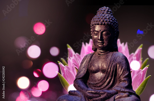 Buddha statue sitting in meditation pose against deep dark background and pink lotus flower behind.