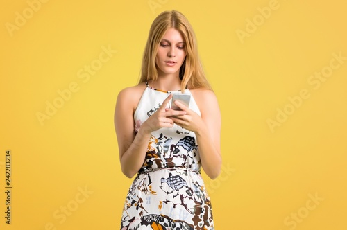 Blonde young girl in a summer dress sending a message or email with the mobile on yellow background