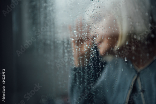 blurred portrait of lonely senior woman at home through window with raindrops