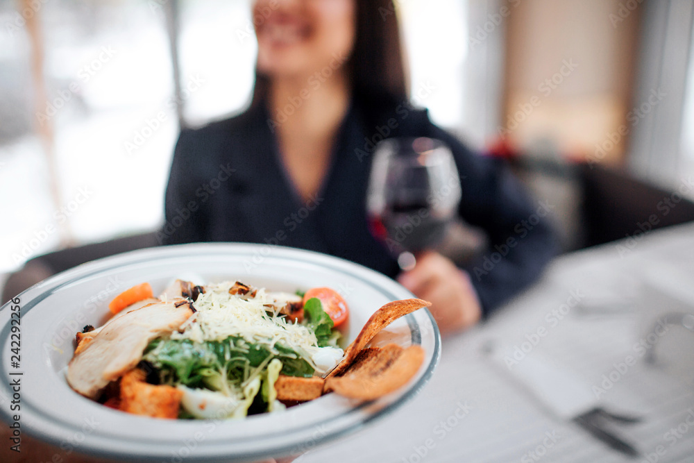 Cheerful young female customer sit at table and smile. She hold glass of red wine. Bowl with salad and meat shown on picture.