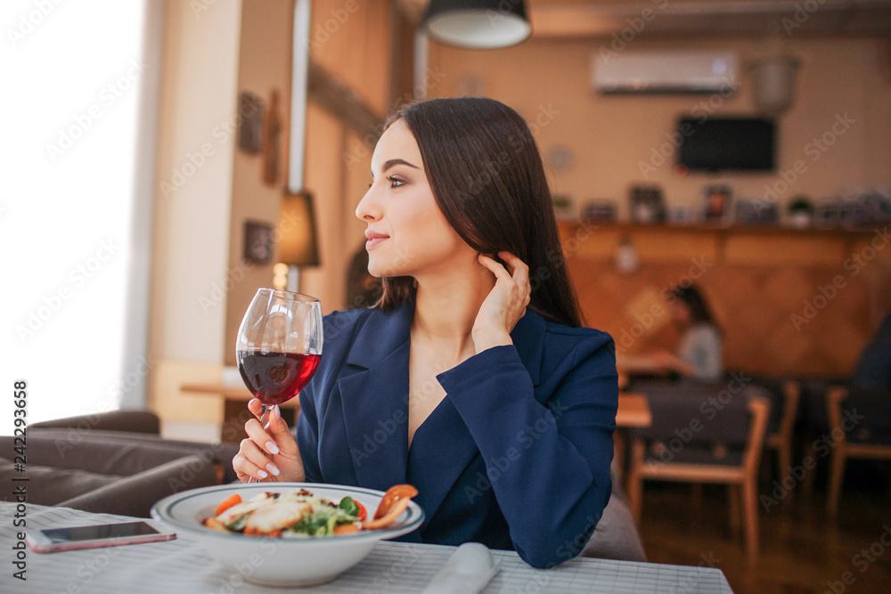 Beautiful young woman sit at table and look to left. She hold glass of red wine and touch her hair. Salad bown stand in front of her. Phone lying besides.