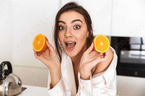 Photo of positive woman 30s wearing silk clothing holding two pieces of orange, during breakfast in kitchen at home