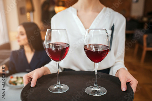 Cut view of waitress hold two glasses of red wine on tray. Youn businesswoman sit behind her at table and look left. She has salad bowl on table.