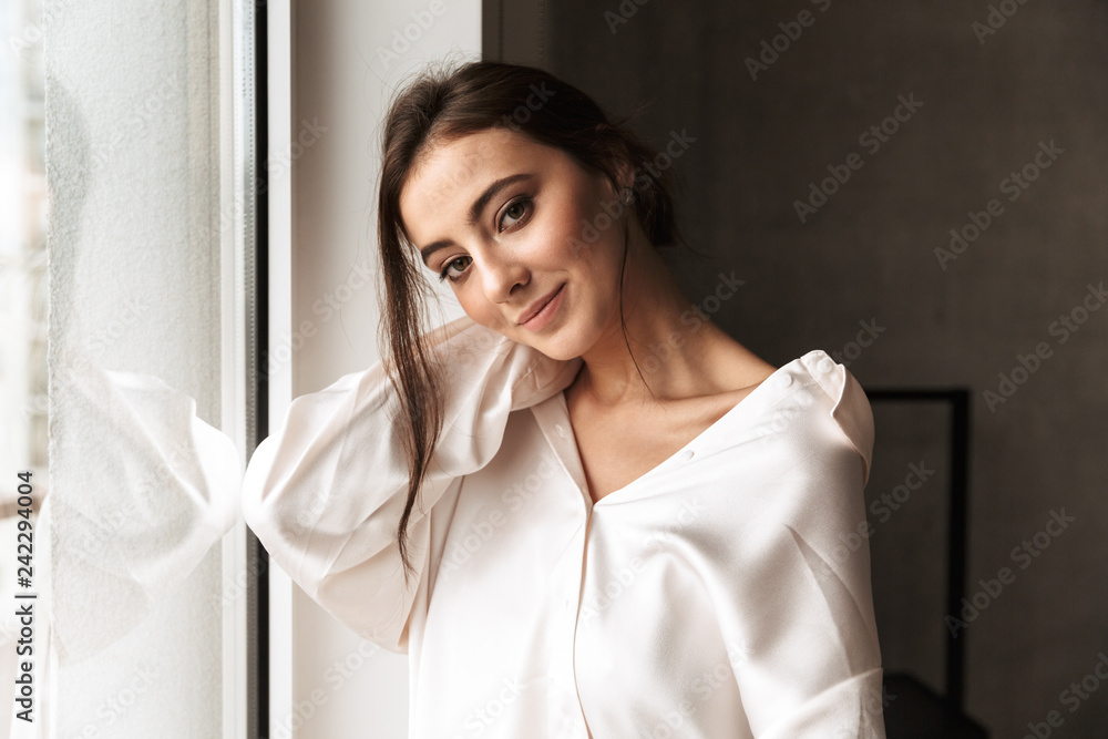 Image of gorgeous woman 30s smiling, and standing near big window in living room Image of gorgeous woman 30s smiling, and standing near big window in living room