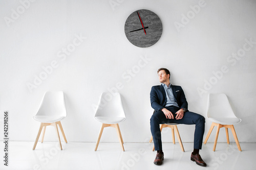 Young man in suit sit on white chair in white room. He lean to wall. Guy wait fo flight in waiting room. He sits alone.