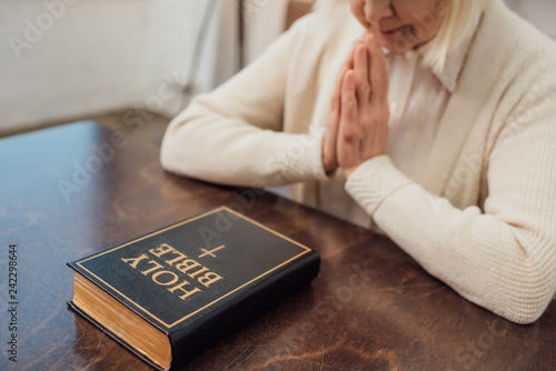 partial view of senior woman sitting and praying in front of holy bible at home