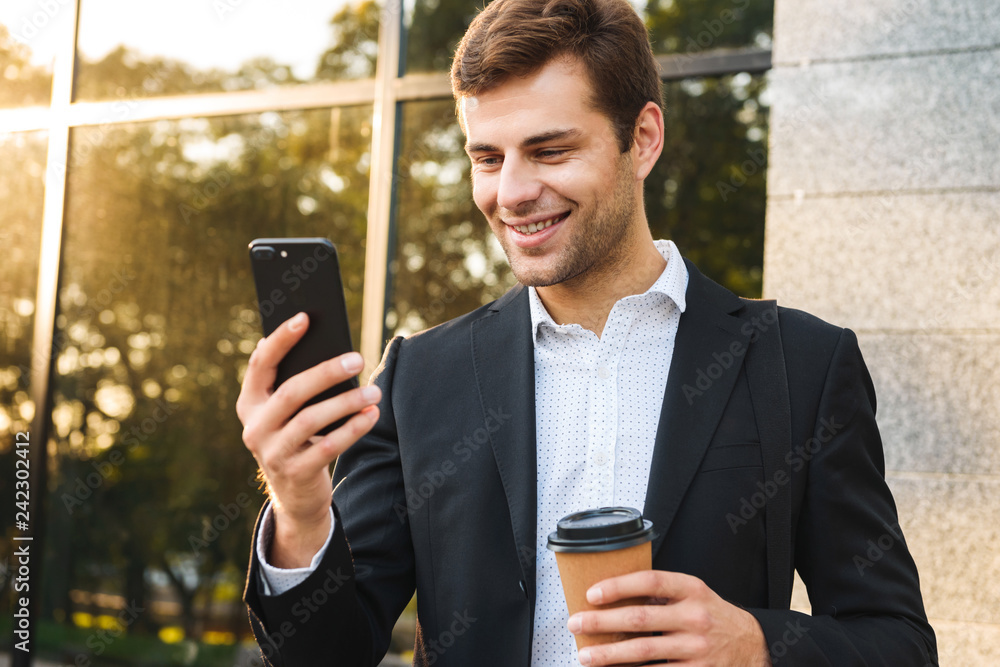 Portrait of executive businessman in suit holding cell phone, while standing outdoor near building with takeaway coffee Portrait of executive businessman in suit holding cell phone, while standing outdoor near building with takeaway coffee