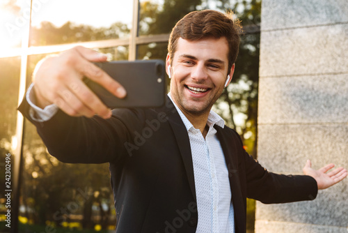 Photo of smiling office worker in suit holding mobile phone for selfie, while standing outdoor against building