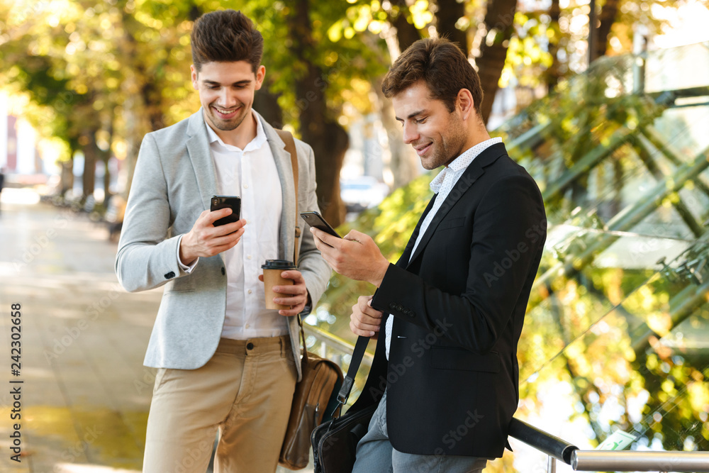 Portrait of businesslike men in suits using smartphone while walking outdoor through green park with takeaway coffee, during sunny day Portrait of businesslike men in suits using smartphone while walking outdoor through green park with takeaway coffee, during sunny day