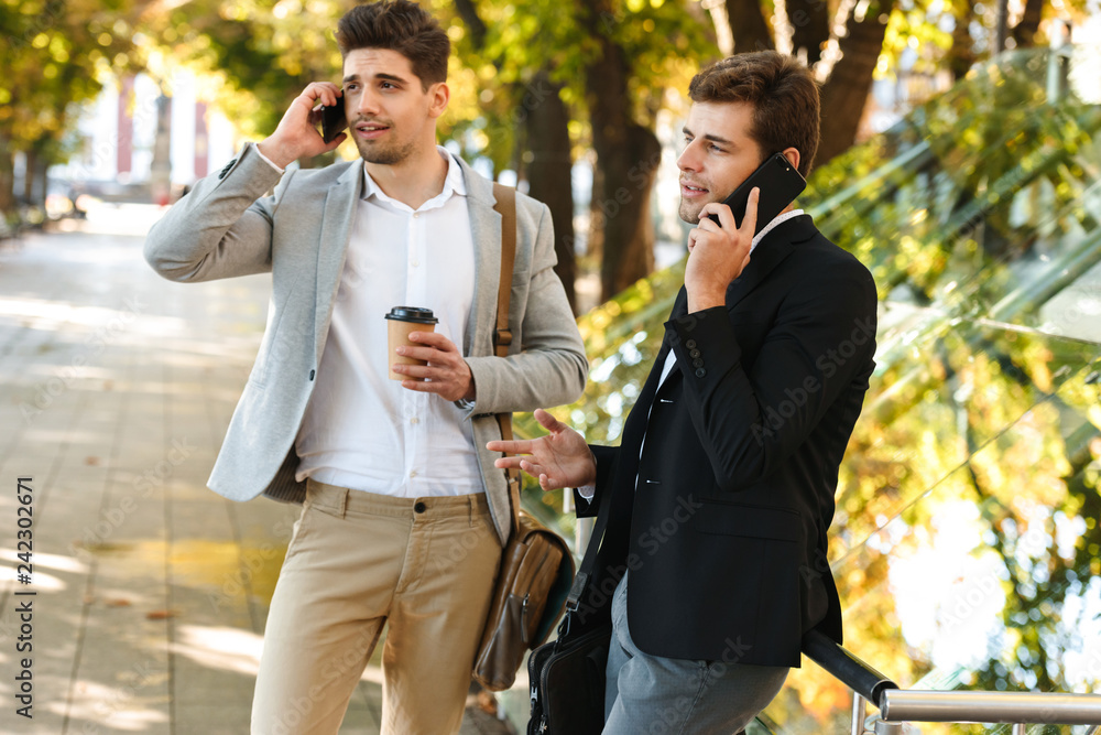 Photo of modern businessmen in suits using smartphone while walking outdoor through green park with takeaway coffee, during sunny day Photo of modern businessmen in suits using smartphone while walking outdoor through green park with takeaway coffee, during sunny day