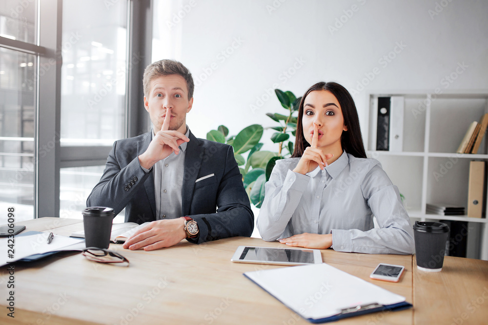 Attentive young man and woman sit at table and look on camera. They are in meeting room. People hold finger on lips. This is hush sign.