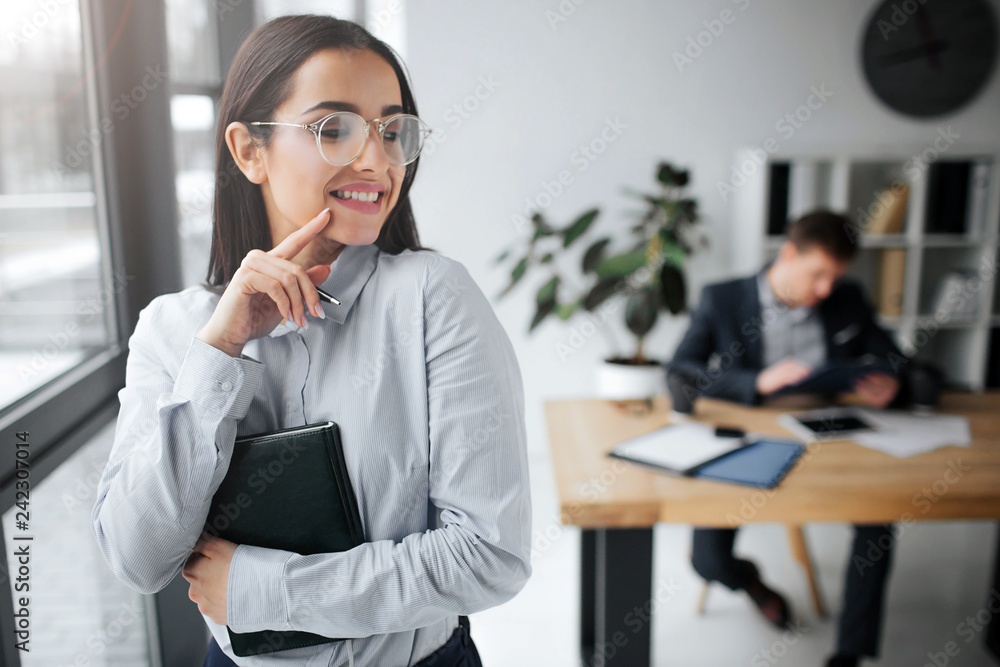 Cheerful and positive young woman stand and smile. She touch her hair and look back at boss. He sit at table. Guy look at brunette. Cheerful and positive young woman stand and smile. She touch her hair and look back at boss. He sit at table. Guy look at brunette.