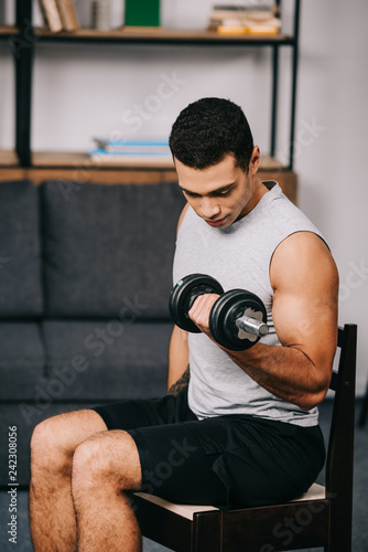 strong mixed race man training in sportswear with heavy dumbbell on chair