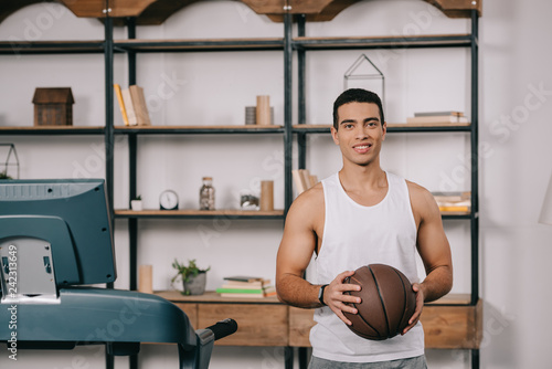 Fototapeta Naklejka Na Ścianę i Meble -  smiling mixed race man holding basketball in living room