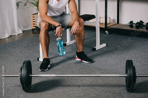 cropped view of man sitting near barbell and holding sport bottle