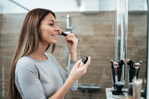 Fotografie Smiling young woman applying make-up in the bathroom, side view.