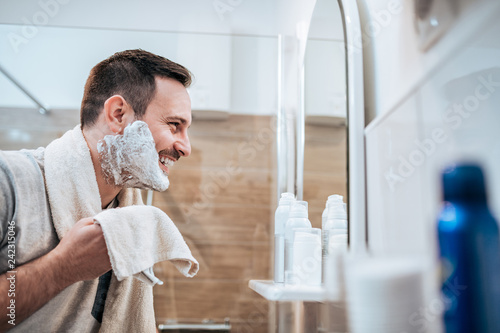 Portrait of a handsome man with shaving cream on his face and towel while standing in the bathroom, looking at mirror. photo