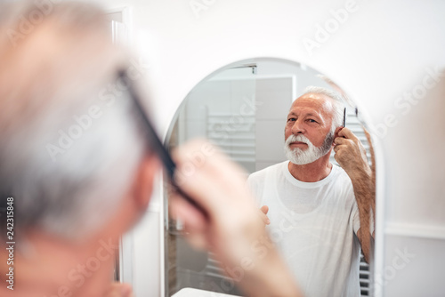 Handsome senior man combing his hair in the bathroom. Focus on the reflection in the mirror.