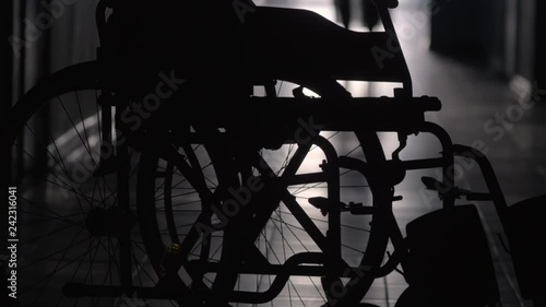 Tilt up shot of silhouette of woman with stick limping to wheelchair along hallway of hospital