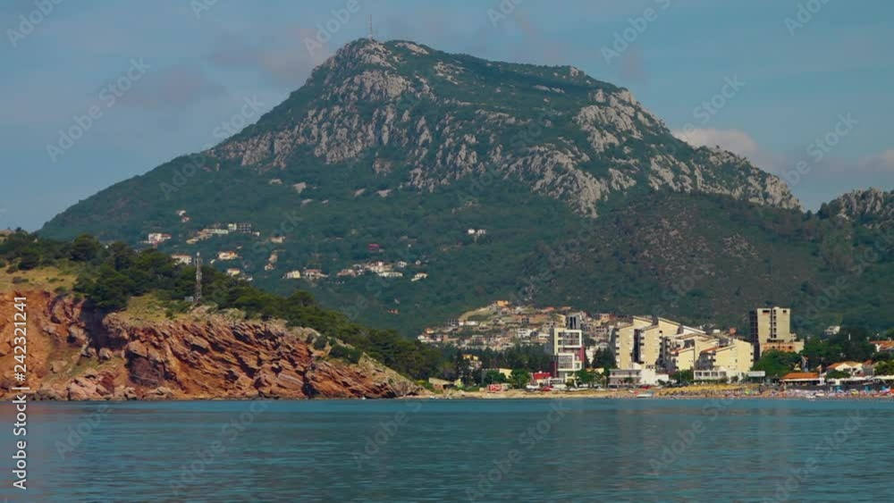 Mid shot calm ocean and a rugged mountain covered in vegetation with a beach and buildings 