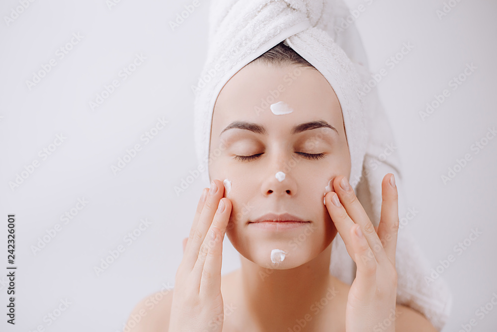 A young beautiful girl after a shower with a towel on her head apply a cleansing mask on her face. Skin care and beauty concept. Place for text