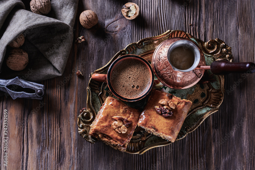A cup of freshly brewed coffee next to a cezve and baklava on a wooden vintage tray and on a wooden background next to nuts, top view, closeup A cup of freshly brewed coffee next to a cezve and baklava on a wooden vintage tray and on a wooden background next to nuts, top view, closeup