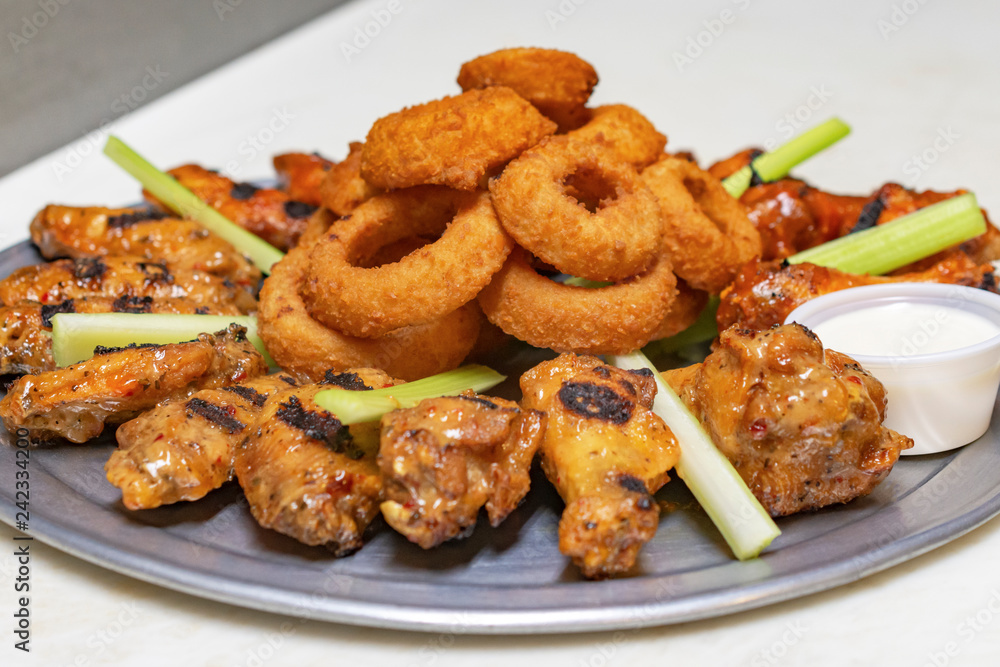 Bar Food:  Chicken wing platter with onion rings and celery.  Container of white dipping sauce on white board.  Side view close up.