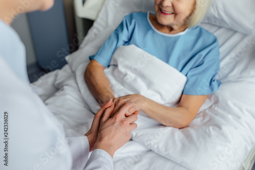 cropped view of female doctor holding hands with senior woman lying in bed in hospital