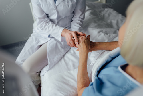 partial view of female doctor holding hands with senior woman lying in bed in hospital