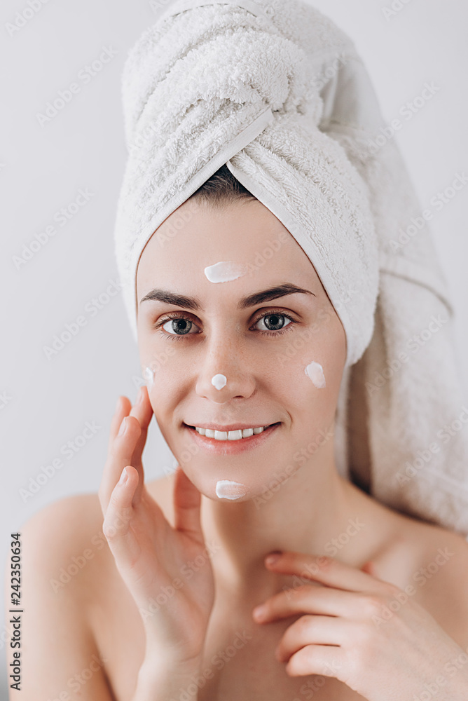 Young happy woman wrapped with bath towel applying cream to her face for skin care. Skin care concept. Stunning caucasian woman with perfect health clean skin. Young happy woman wrapped with bath towel applying cream to her face for skin care. Skin care concept. Stunning caucasian woman with perfect health clean skin.