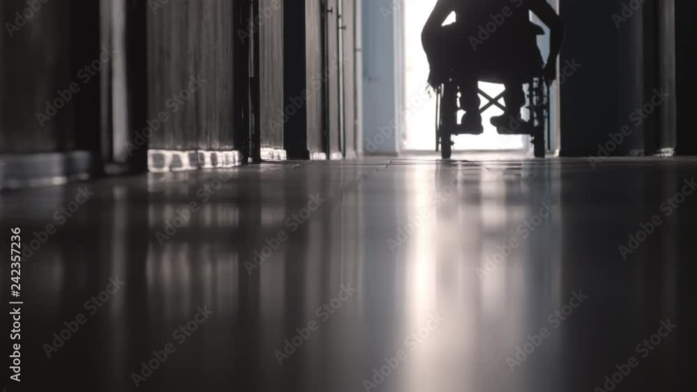 Low section of silhouette of disabled man riding wheelchair towards the camera along hospital hallway Low section of silhouette of disabled man riding wheelchair towards the camera along hospital hallway