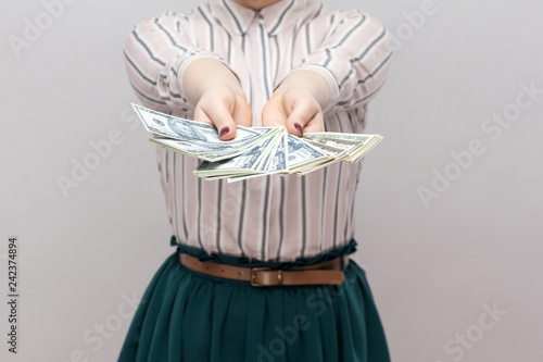 Take it! Close up portrait of successful woman in striped shirt standing, holding many dollars, demonstration fan of money. Indoor, studio shot, isolated, gray background
