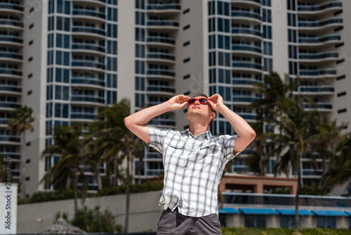 Young man hipster millennial looking up on beach during sunny day with in Miami Florida with hands on sunglasses watching apartment condo skyscraper