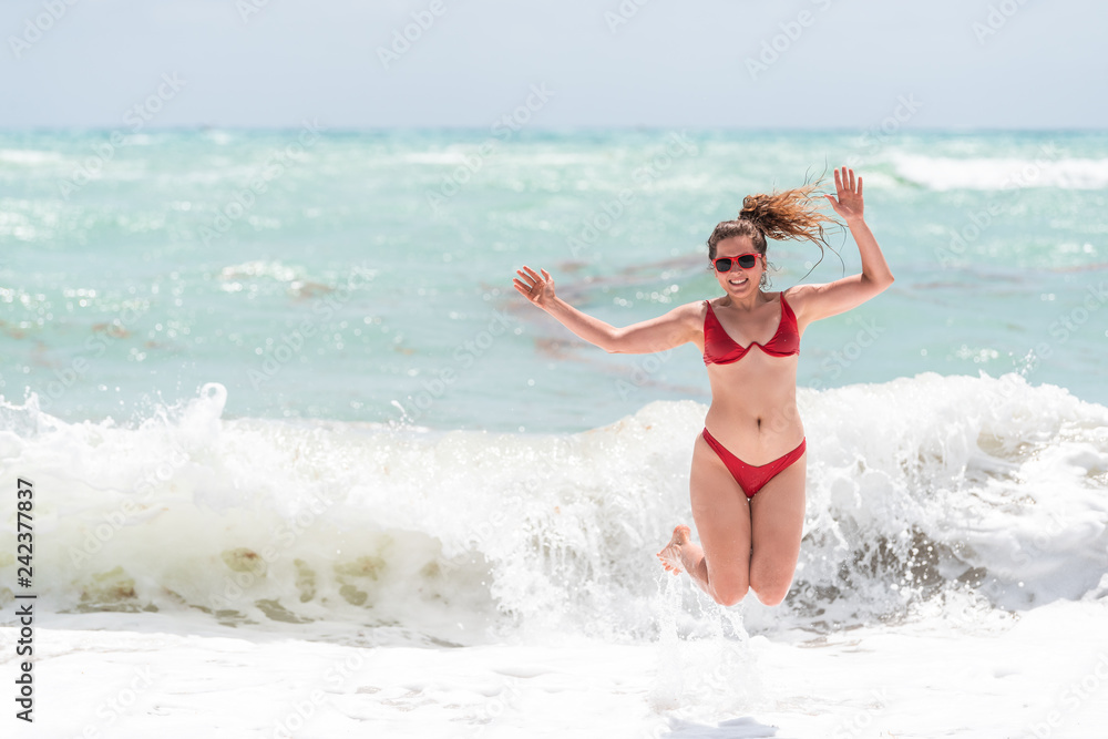Young happy woman jumping up with arms raised waving in red swimsuit bikini bathing suit and splash crashing wave in Miami Beach Florida green water drops Young happy woman jumping up with arms raised waving in red swimsuit bikini bathing suit and splash crashing wave in Miami Beach Florida green water drops