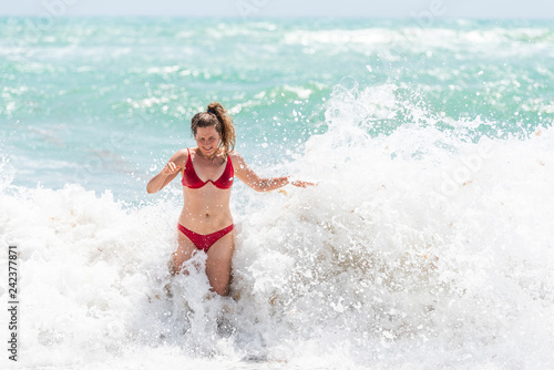 Young woman standing in red swimsuit bikini bathing suit scared surprised by splash crashing wave in Hollywood, Miami Beach Florida green water drops