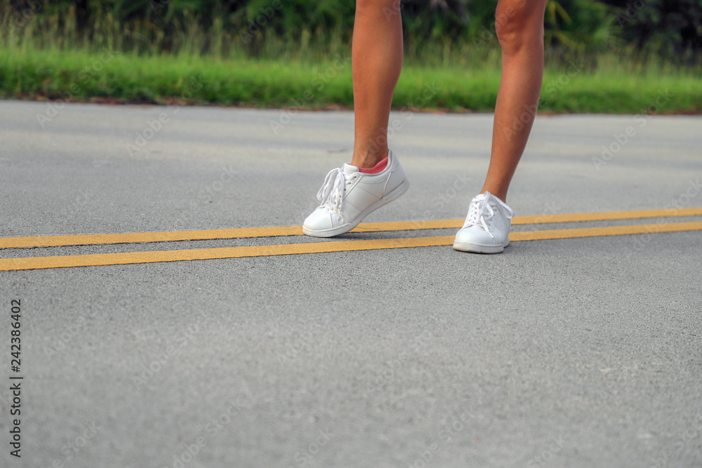 Tanned sporty female legs in white sneakers on the asphalt road on the yellow strip Tanned sporty female legs in white sneakers on the asphalt road on the yellow strip