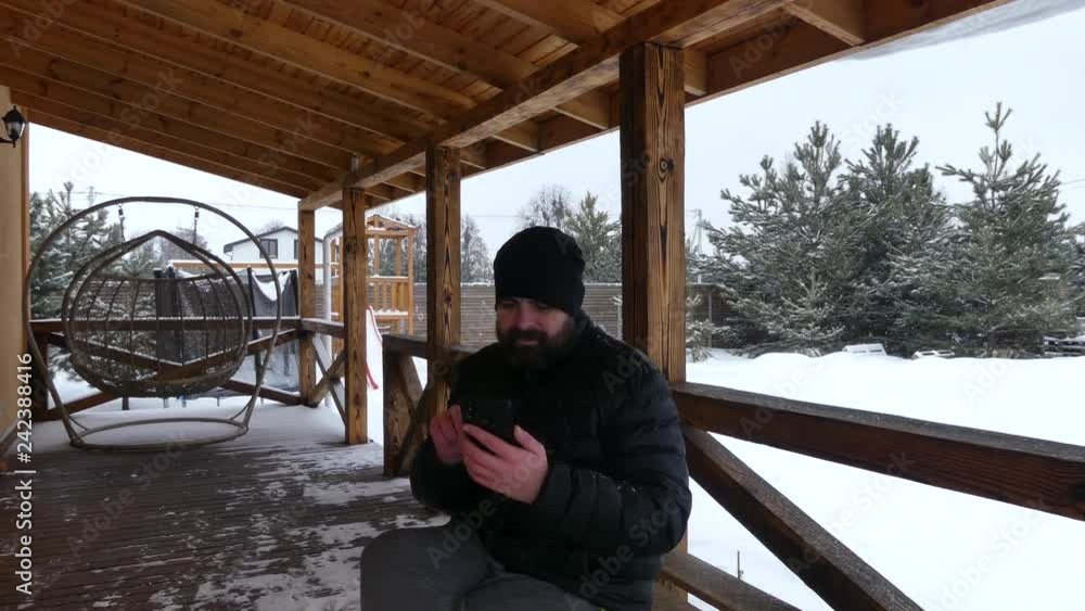 Bearded man smokes a cigarette and looks into the smartphone on the wooden terrace of the cottage. Wooden terrace in the winter among the pines. Bearded man smokes a cigarette and looks into the smartphone on the wooden terrace of the cottage. Wooden terrace in the winter among the pines.