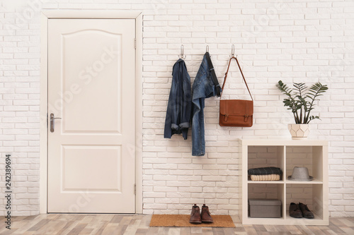 Stylish hallway interior with door, comfortable furniture and clothes on brick wall