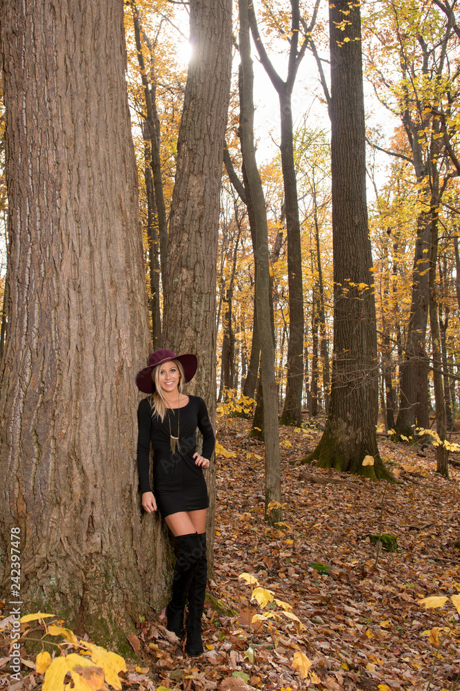 Sexy young blonde woman in stunning black dress and over the knee boots  wears a colorful hat in the autumn woods - fall fashion - sun shining  through trees above model foto