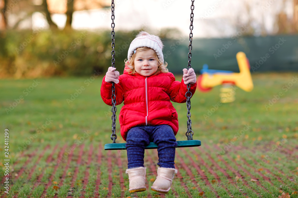 Cute toddler girl having fun on playground. Happy healthy little child climbing, swinging and sliding on different equipment. On cold day in colorful clothes. Active outdoors game for children